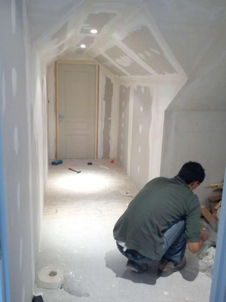 Personne agenouillée, en train de cloisonner un couloir avec un plafond en pente et une porte fermée en arrière-plan.