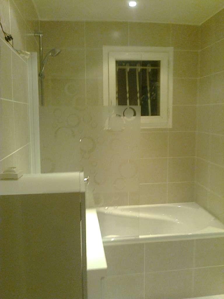 Salle de bain carrelée beige avec baignoire blanche, fenêtre avec barreaux et pommeau de douche.