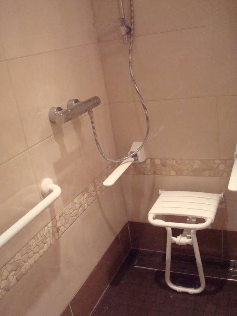 Cabine de douche avec siège rabattable blanc et barre d'appui, murs carrelés beiges et pommeau de douche à main.