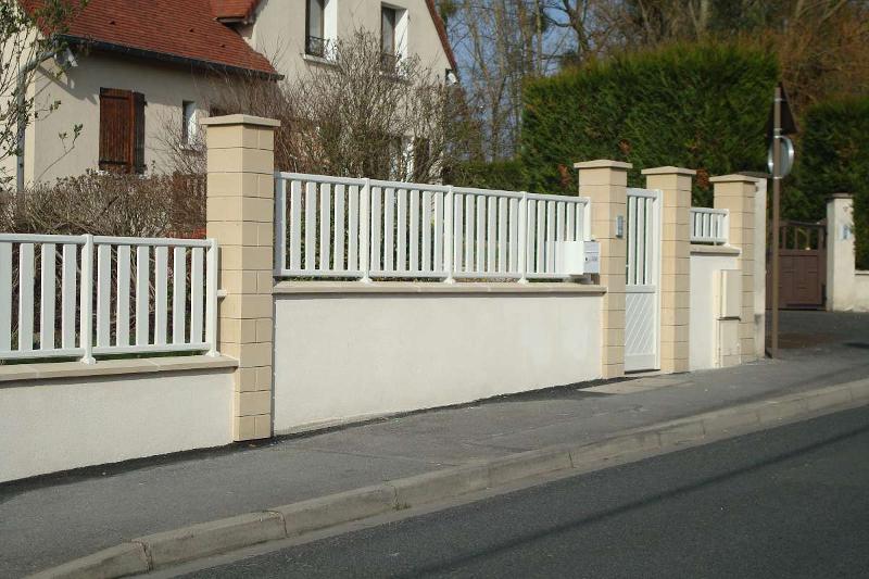 Clôture blanche et portail devant un mur beige clair, situé le long d'une rue, avec une maison et des arbres en arrière-plan.