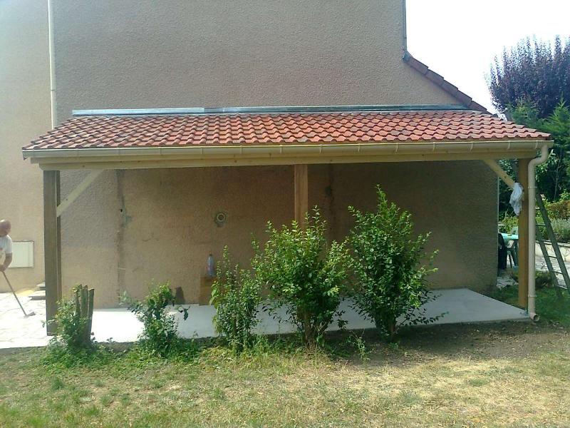 Terrasse couverte en bois accolée à un bâtiment beige au toit de tuiles rouges ; buissons au premier plan, pelouse devant.