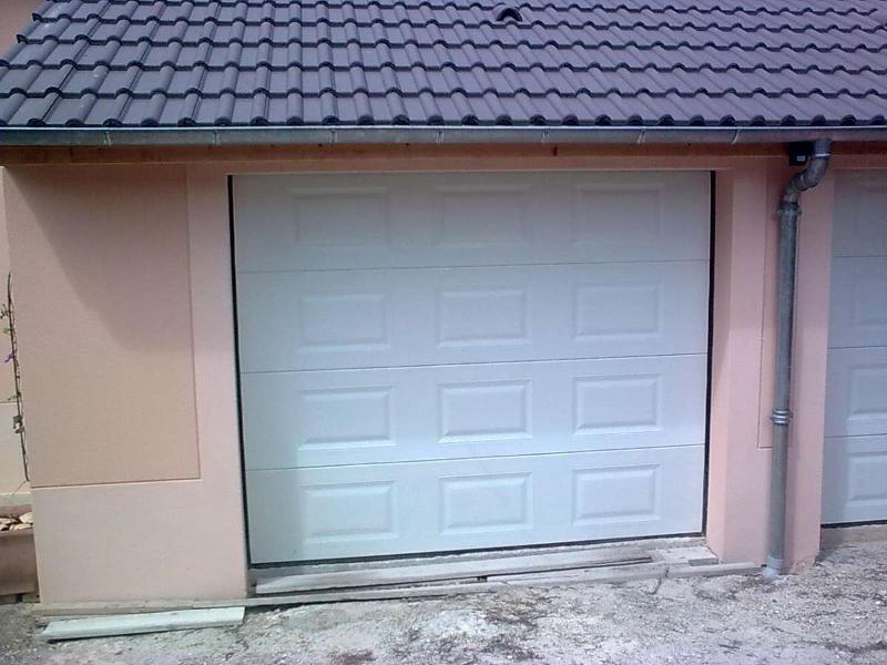 Porte de garage blanche sur un bâtiment rose avec un toit gris et une descente pluviale.