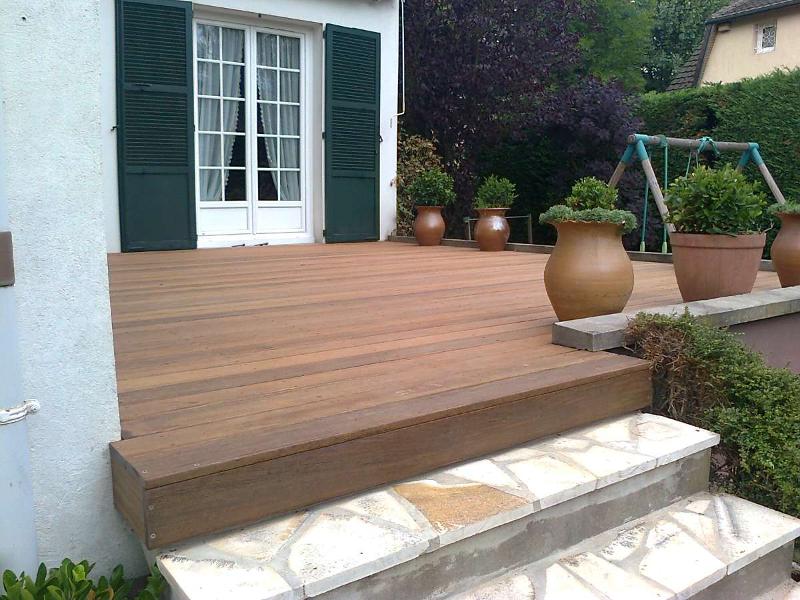 Terrasse en bois avec marches en pierre menant à une maison blanche avec des volets verts et des plantes en pot.