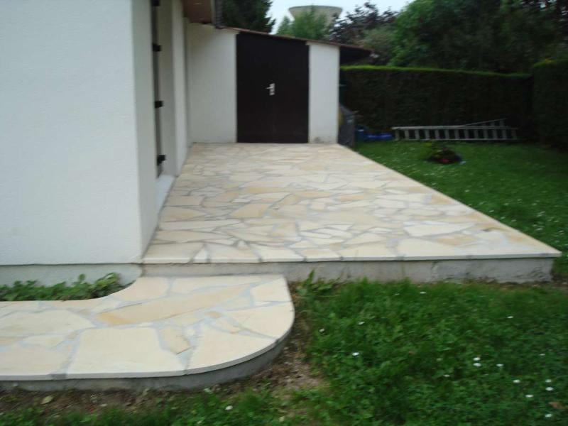 Terrasse en pierre à côté d'un bâtiment blanc et d'un hangar brun foncé. Autour, une pelouse verte.