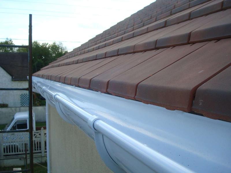 Gouttière blanche le long d'une ligne de toit en tuiles de terre cuite, à côté d'une maison beige avec un véhicule visible.