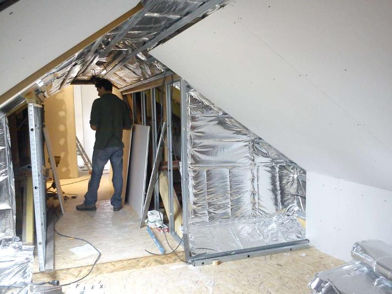 Homme debout dans un grenier en cours de rénovation, charpente métallique, isolation réfléchissante, cloison sèche.