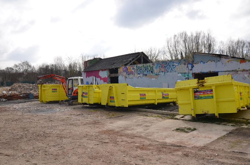Des bennes à ordures jaunes à côté d'un bâtiment couvert de graffitis, sur un terrain gravillonné