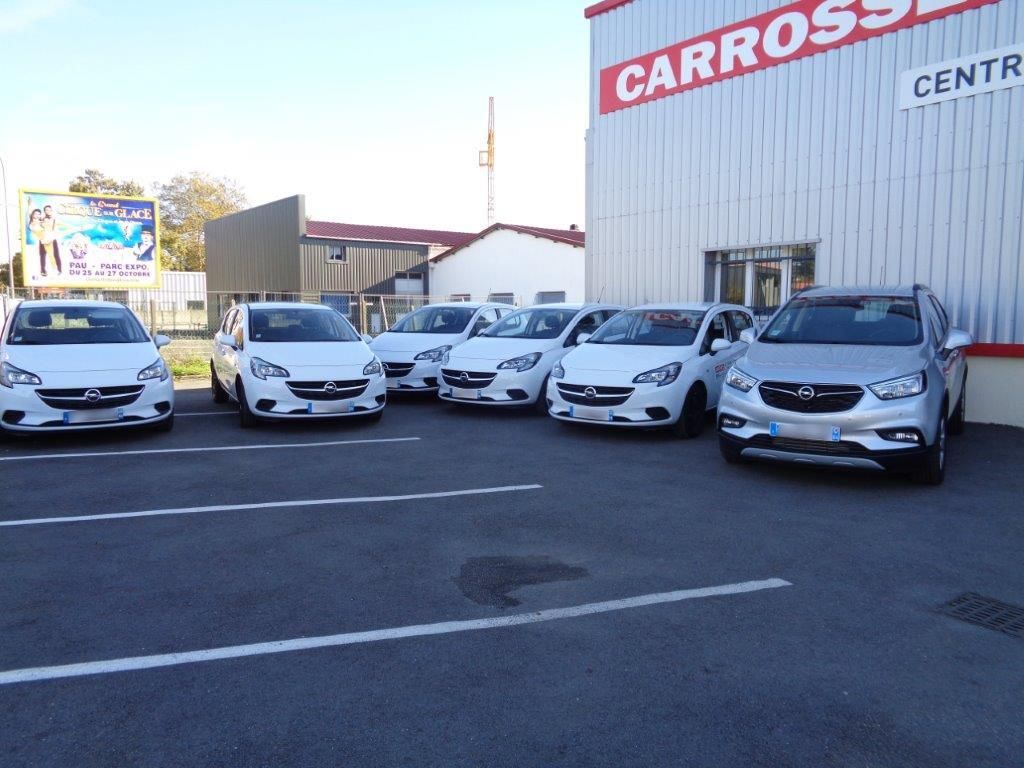 Des voitures blanches garées devant un bâtiment où l'on peut lire le mot CARROSSERIE en lettres rouges.