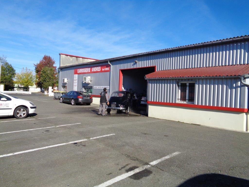Extérieur du garage ; une personne travaille sur une voiture. Parking devant, bâtiment en métal avec des finitions rouges.