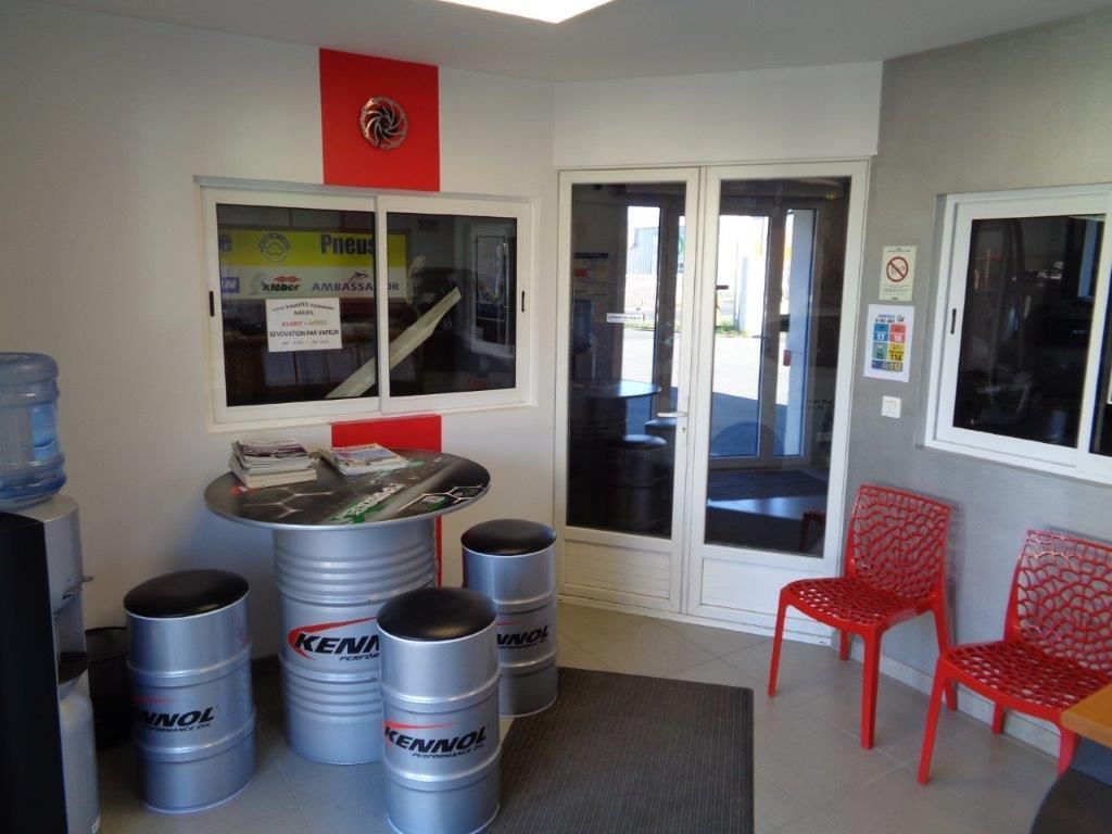 Salle d'attente avec table ronde, tabourets, chaises rouges et double porte. Le logo de l'entreprise est visible.