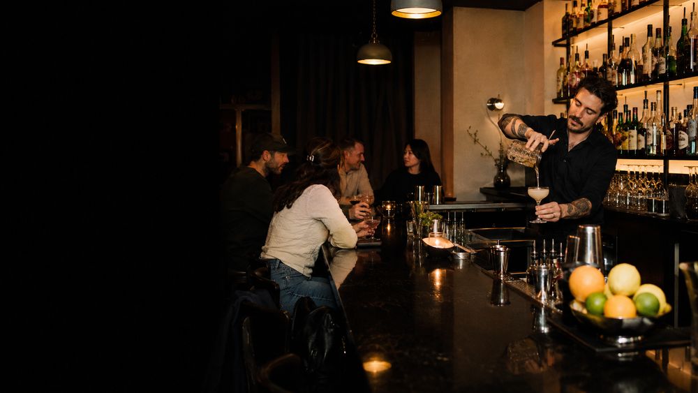 Bartender pouring a cocktail from a mixing tin while guests lean over the counter watching