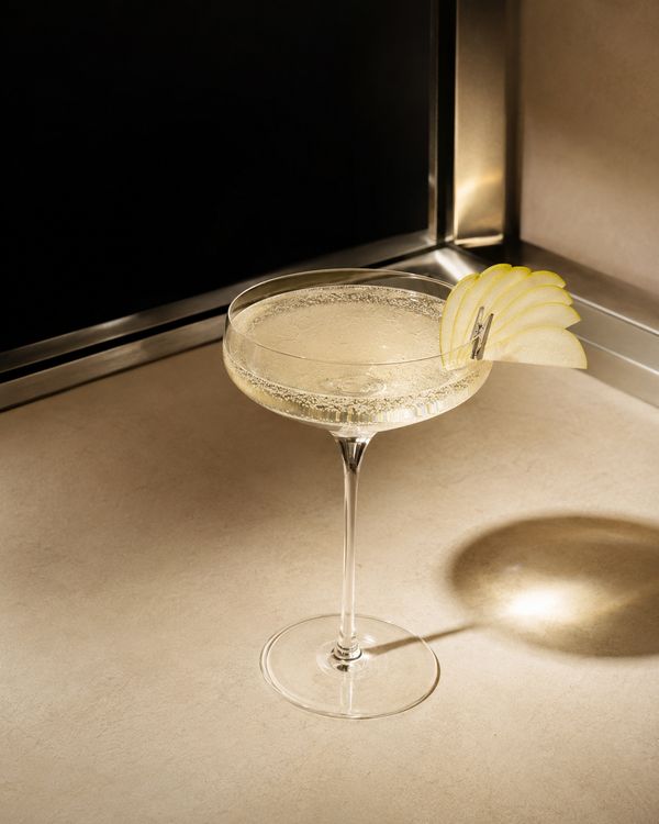 A sparkling pale cocktail in a coupe glass with a thin pear slice resting on the rim