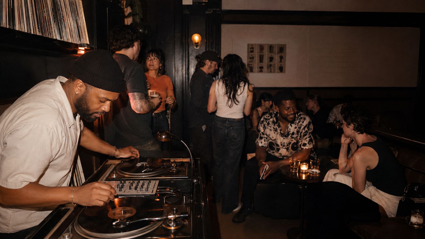A DJ spinning vinyl at a low-lit lounge booth while guests dance and talk nearby