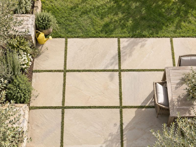 Drone shot straight down at 12m over a new sandstone terrace with grass joints and a single yellow watering can at the edge, soft morning light, clean realistic colour.